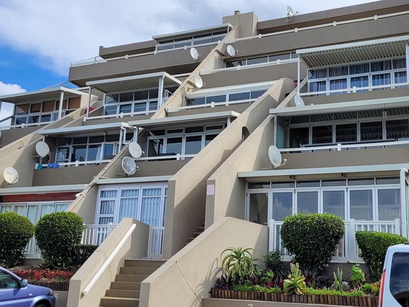 Front elevation of a multi-story apartment complex featuring tiered balconies, concrete architectural detailing, and landscaped garden beds at the entrance.