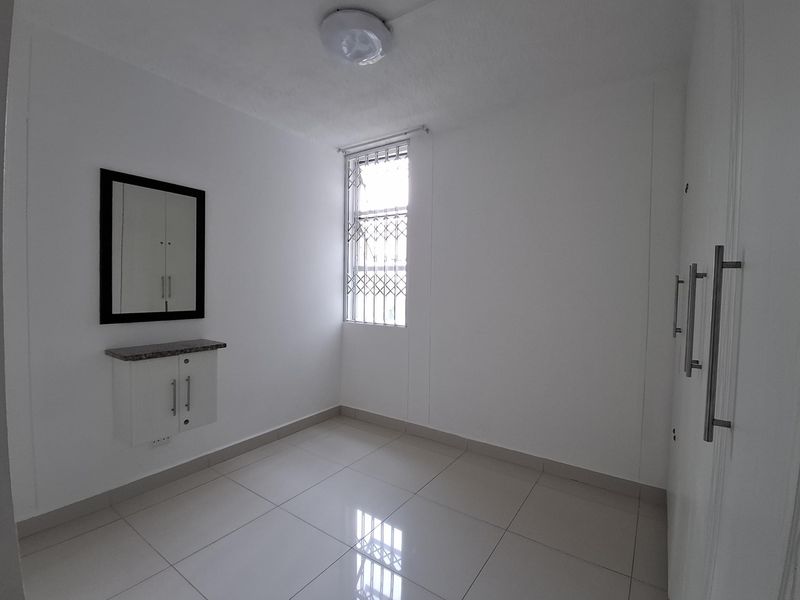 A clean, empty bedroom featuring tiled flooring, built-in cupboards, and a wall-mounted vanity unit with a window fitted with a security gate.