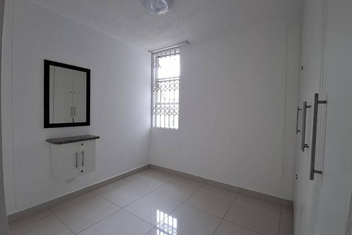 A clean, empty bedroom featuring tiled flooring, built-in cupboards, and a wall-mounted vanity unit with a window fitted with a security gate.