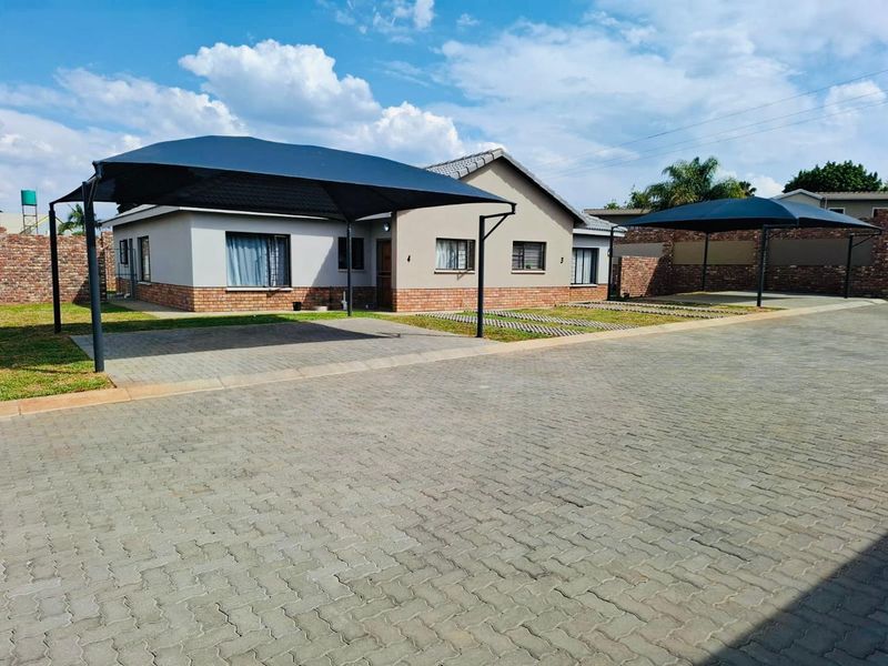 Front exterior view of a townhouse featuring a paved driveway, shade cloth carports, and a well-maintained facebrick and plaster facade.