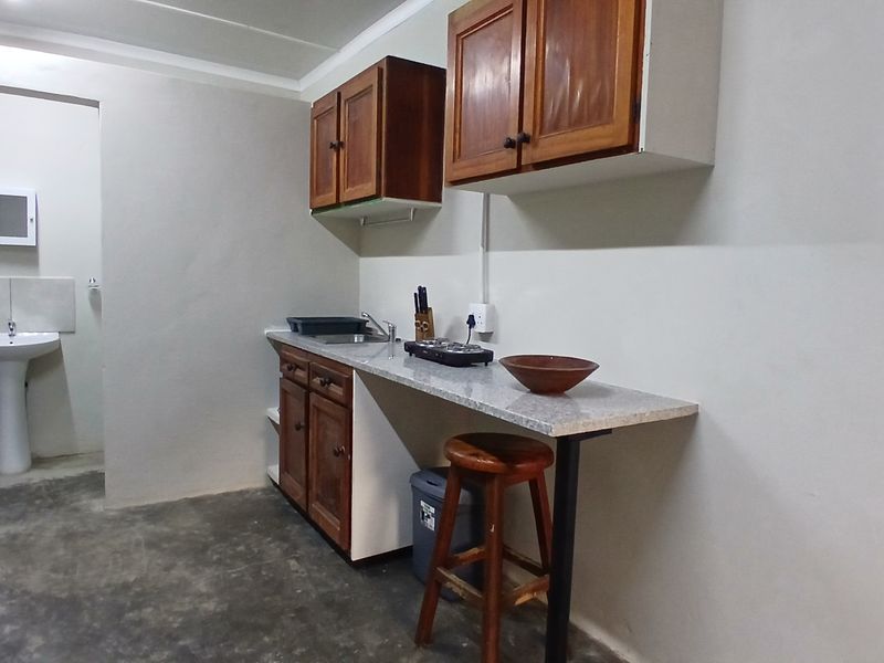 A compact kitchenette featuring wooden cabinetry, a laminate countertop, and an adjacent pedestal basin area with basic finishes.