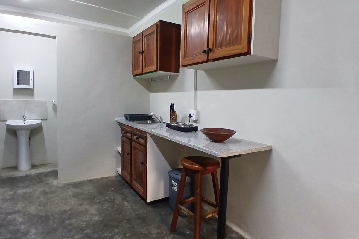 A compact kitchenette featuring wooden cabinetry, a laminate countertop, and an adjacent pedestal basin area with basic finishes.
