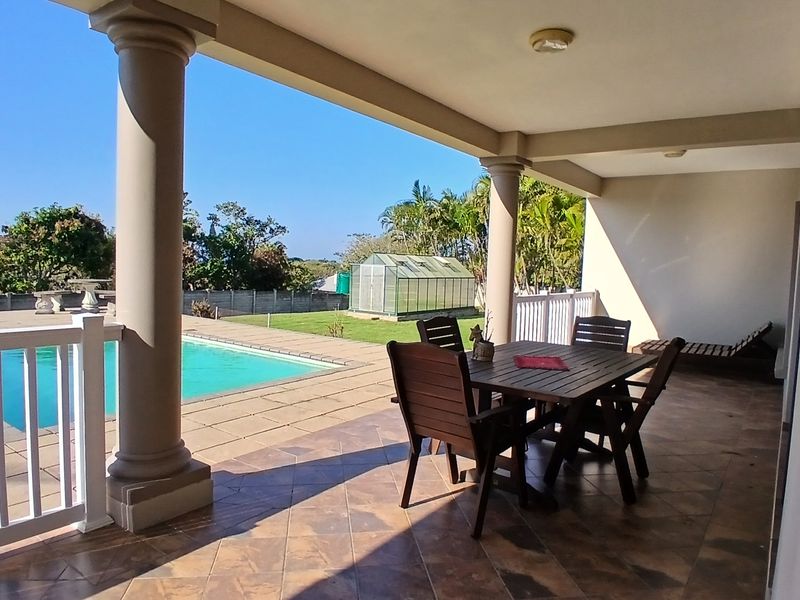 A spacious covered patio featuring tiled flooring and structural columns, overlooking a private swimming pool and garden area.