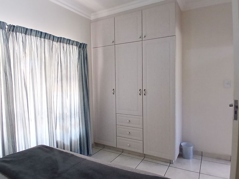 A bedroom featuring built-in cupboards, tiled flooring, and large windows with curtains, offering a clean and functional living space.