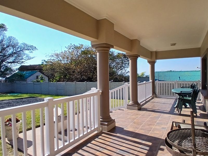 A spacious covered patio featuring tiled flooring, classic columns, and a white balustrade railing, overlooking a private garden area.