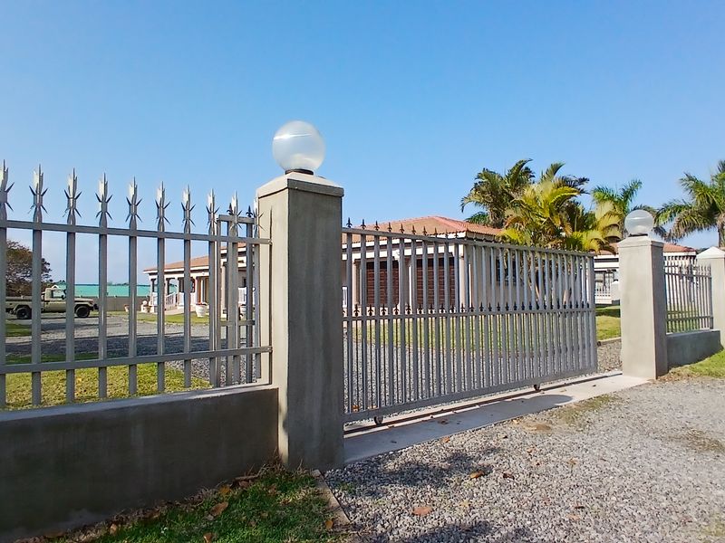 A secure property entrance featuring a metal sliding gate, concrete pillars with integrated lighting, and a gravel driveway leading to the main house.