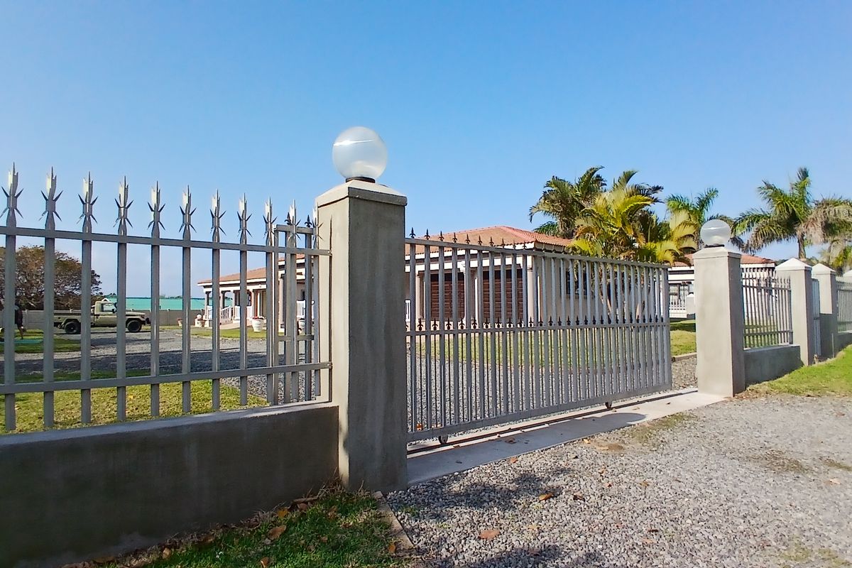 A secure property entrance featuring a metal sliding gate, concrete pillars with integrated lighting, and a gravel driveway leading to the main house.