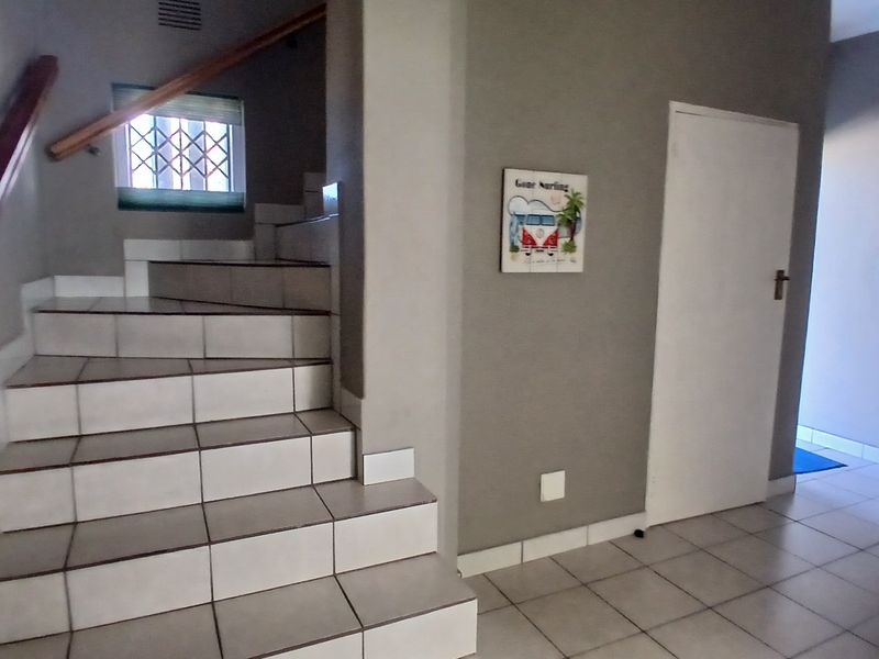 A tiled hallway featuring a staircase with a wooden handrail and a window with security bars, leading to adjacent rooms.