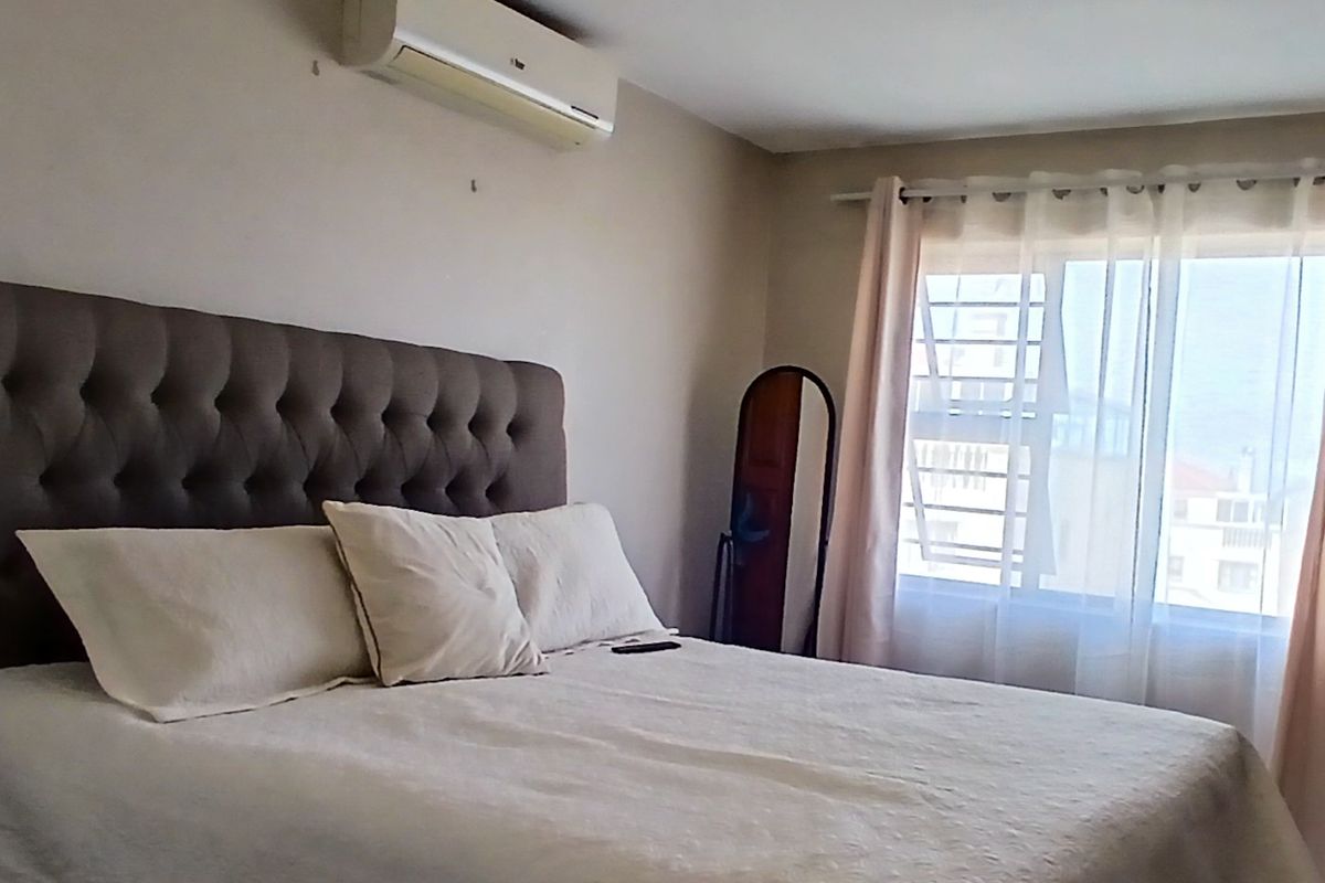 A bedroom featuring a wall-mounted air conditioning unit and a window fitted with security bars, allowing natural light into the space.