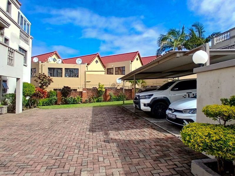 A paved communal courtyard area featuring a covered carport, brick boundary walls with security fencing, and access to the apartment building.