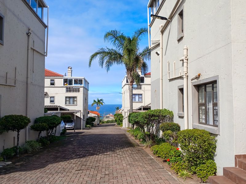 A view down a paved driveway between apartment blocks, featuring external building infrastructure and a glimpse of the ocean in the distance.