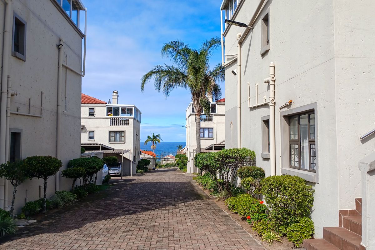 A view down a paved driveway between apartment blocks, featuring external building infrastructure and a glimpse of the ocean in the distance.