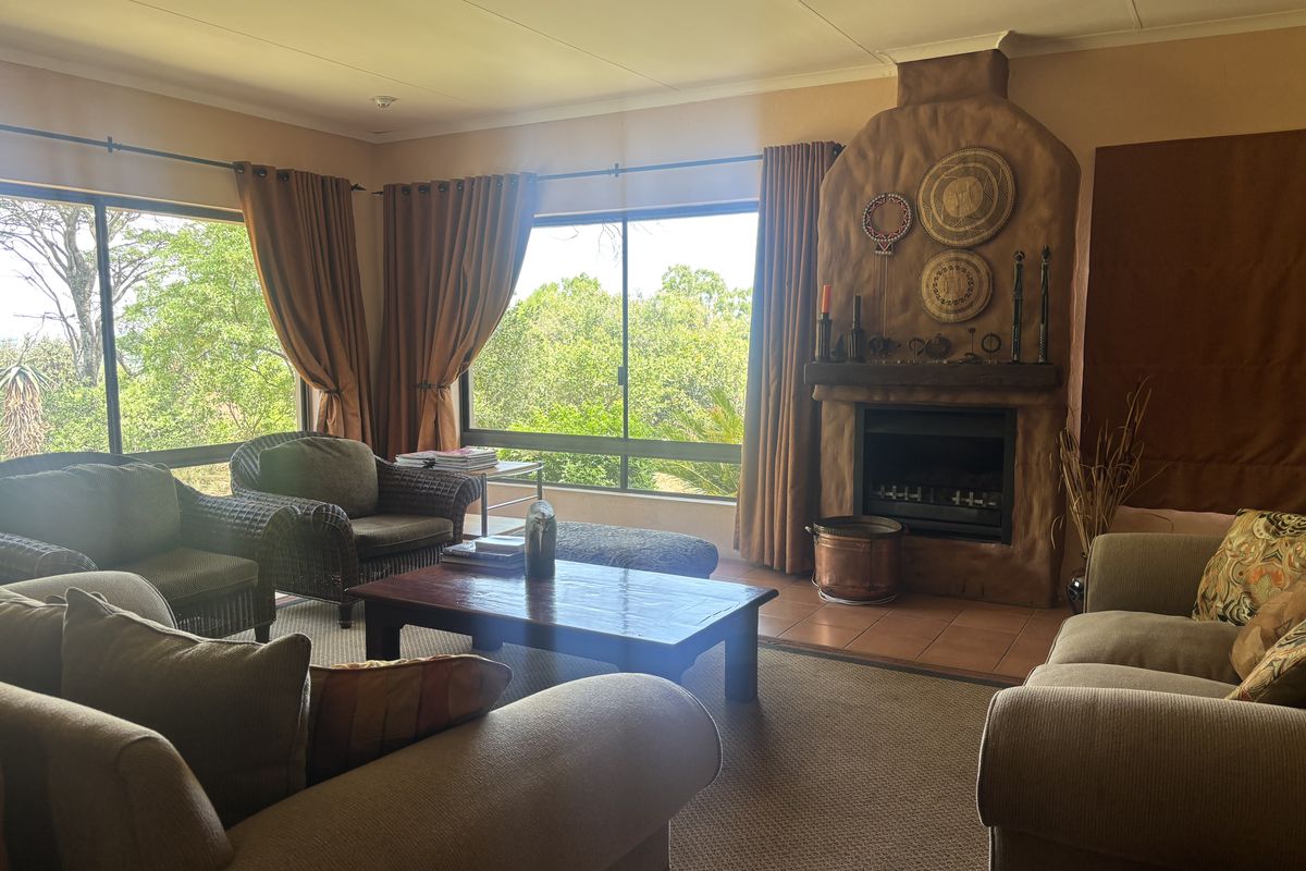 A cozy living room featuring a prominent rustic fireplace, large windows with garden views, and neutral carpeting.