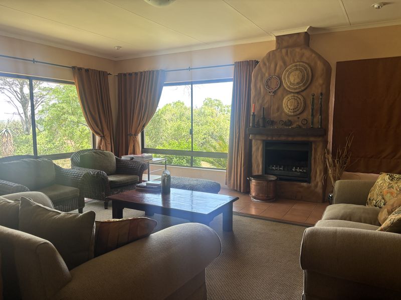 A cozy living room featuring a prominent rustic fireplace, large windows with garden views, and neutral carpeting.
