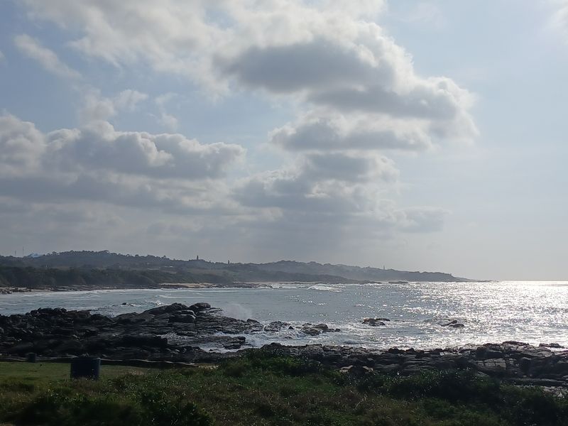 A scenic coastal view featuring rocky shoreline, ocean waves, and a distant landscape under a cloudy sky.