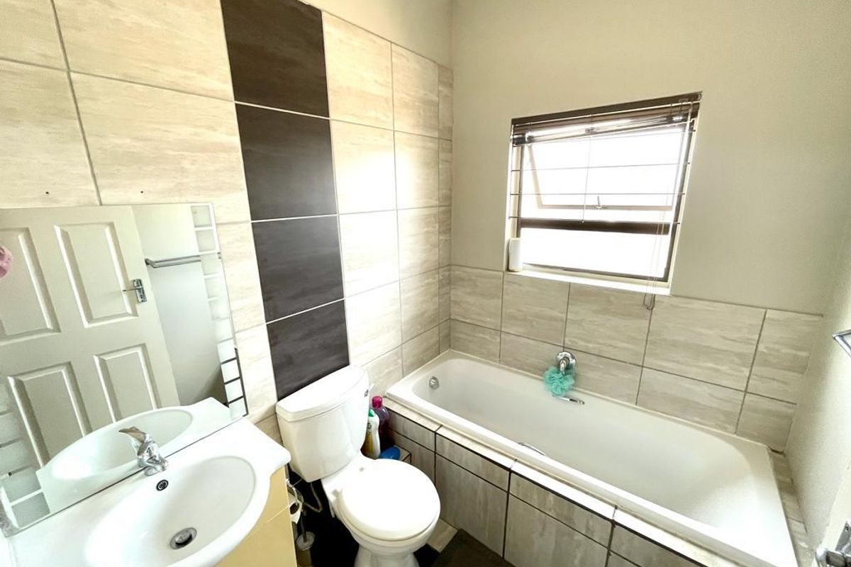 A functional bathroom featuring a built-in bathtub, a vanity basin with a mirror, and a toilet, finished with neutral wall tiling.
