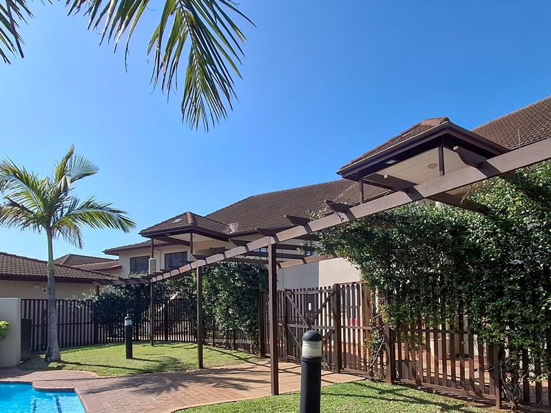 Outdoor pool area featuring a paved patio, timber pergola, and established garden surroundings within a residential complex.