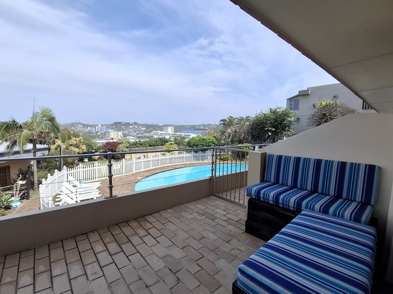 A covered balcony featuring brick paving and metal railings, offering an elevated view overlooking the complex swimming pool and surrounding landscape.
