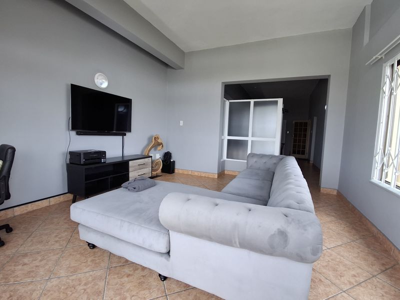 A spacious living room featuring neutral grey walls, tiled flooring, and a large window with security bars, providing access to an adjacent hallway.