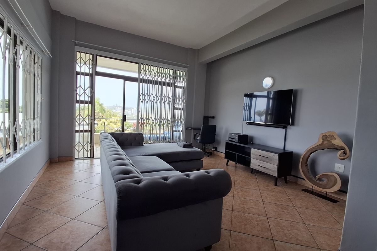 A spacious living room featuring tiled flooring, neutral grey walls, and a large sliding glass door with security gates leading to an outdoor area.