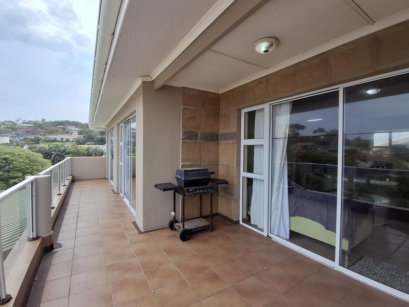 A spacious covered balcony featuring tiled flooring, a modern glass balustrade, and large sliding glass doors providing access to the interior living space.