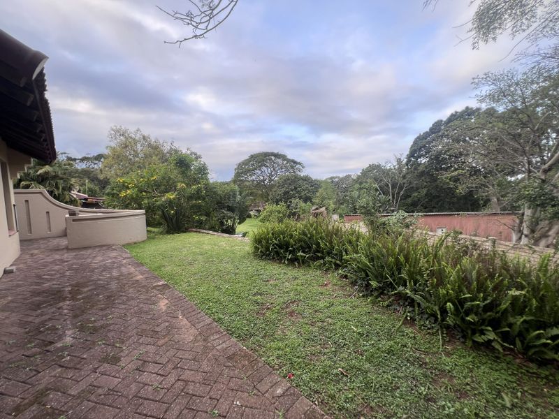 A rear exterior view of the townhouse featuring a paved patio area, a low retaining wall, and a private garden with mature trees.