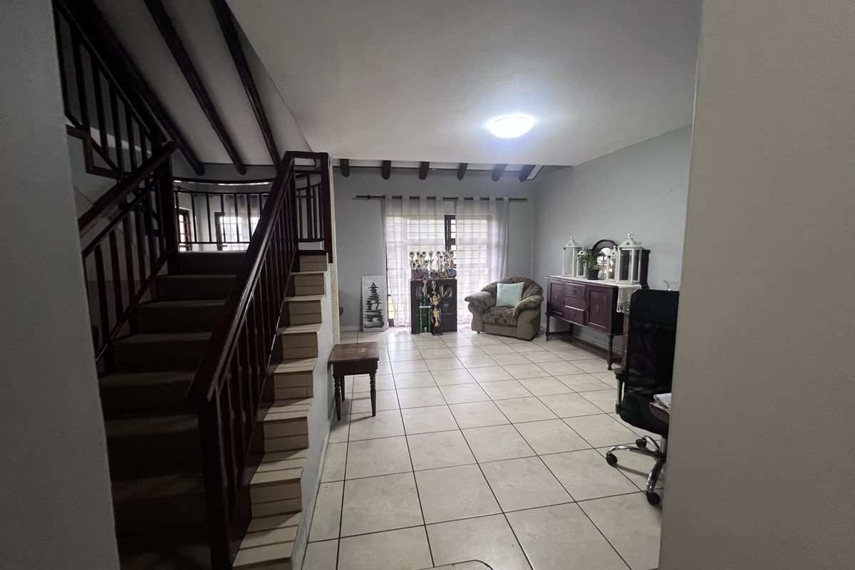 Spacious living area featuring tiled flooring, exposed ceiling beams, and a wooden staircase with a matching balustrade leading to the upper level.