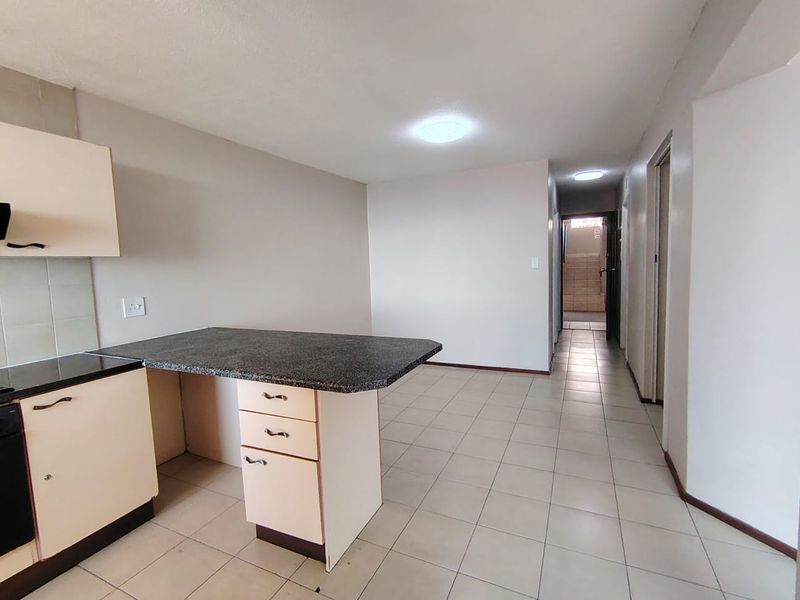 An open-plan kitchen area featuring built-in cabinetry with granite countertops and neutral ceramic floor tiles, leading into a hallway.