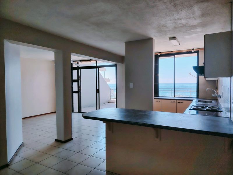 Spacious open-plan living and kitchen area featuring tiled floors, a functional kitchen layout with built-in appliances, and direct access to a balcony with ocean views.