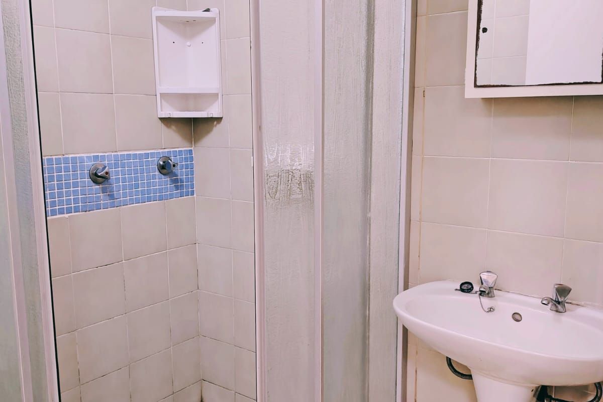 A functional bathroom featuring a shower enclosure with tiled walls, a pedestal basin, and a wall-mounted mirror, requiring some cosmetic updates.