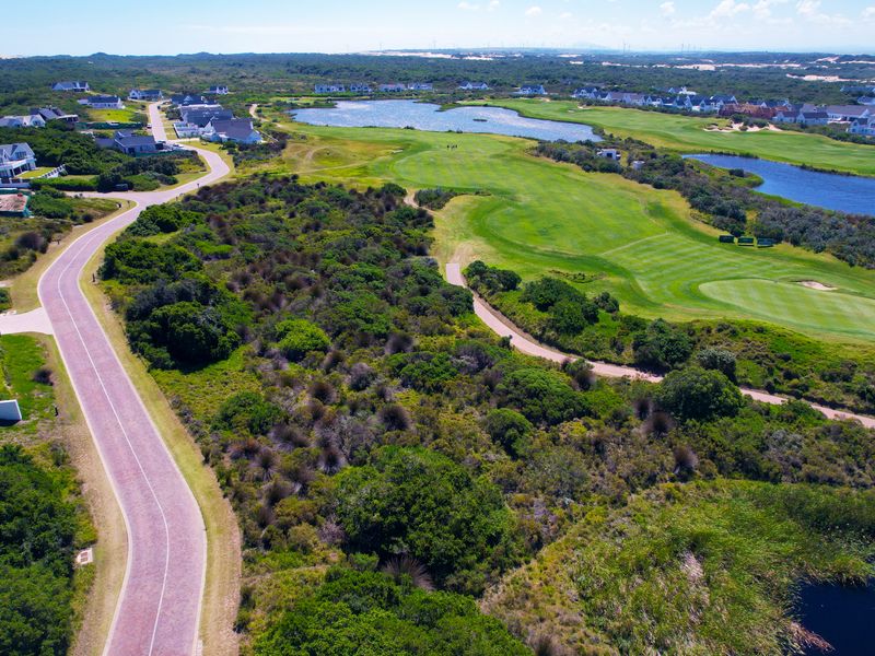 An aerial view of a residential estate featuring a paved road, natural coastal vegetation, and an adjacent golf course with water features.