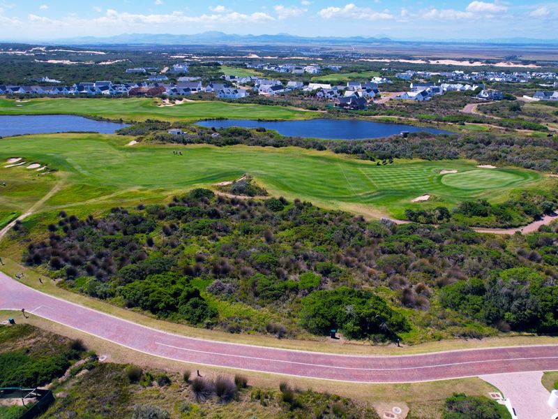 Aerial view of a vacant residential plot situated within a golf estate, featuring surrounding natural vegetation, a paved road, and scenic landscape views.