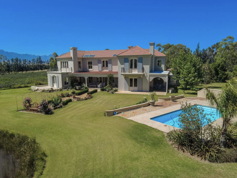 Aerial view of a large farmhouse featuring a swimming pool, expansive manicured lawns, covered patio, and balconies set against a scenic landscape.