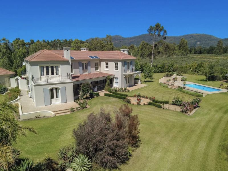 Aerial view of a large farm estate featuring a well-maintained residence, expansive lawns, landscaped gardens, and a private swimming pool set against a mountain backdrop.