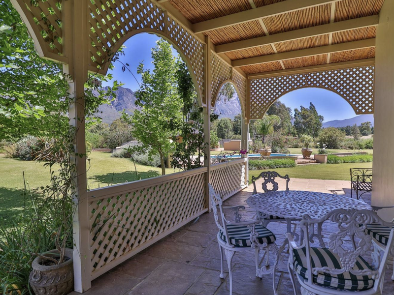 A charming covered patio featuring timber lattice detailing and a thatched roof, overlooking a lush garden with mountain views and a swimming pool.