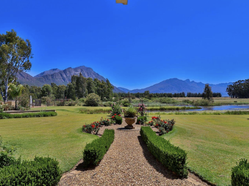 A beautifully maintained farm garden featuring a gravel pathway, manicured hedging, and a serene pond with expansive mountain views.