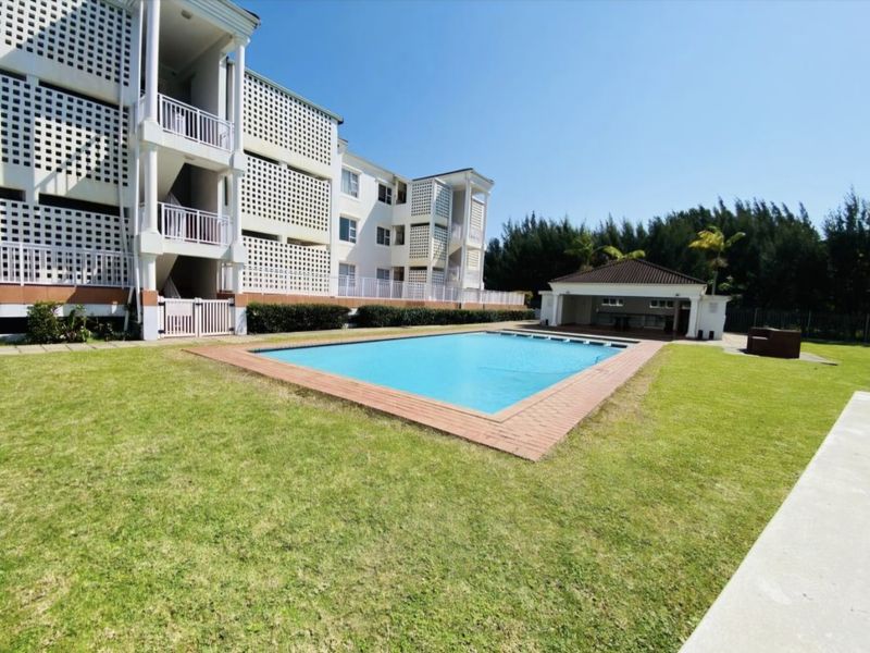 A well-maintained communal swimming pool area featuring a brick-edged pool, landscaped lawn, and a covered poolside pavilion set against the apartment complex.