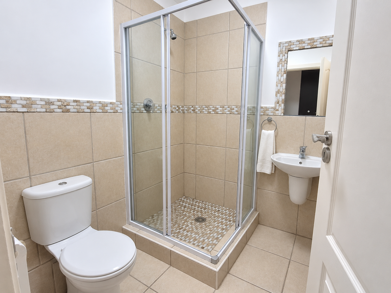 A clean and functional bathroom featuring a corner shower enclosure, wall-mounted basin, toilet, and neutral tiled finishes with a decorative mosaic border.