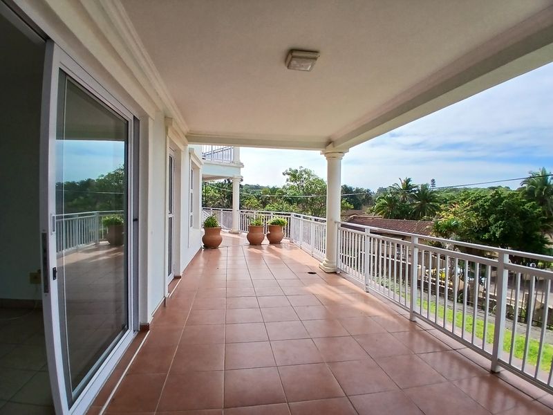 A spacious covered balcony featuring tiled flooring, white railings, and sliding glass door access, offering an open outdoor area with scenic views.