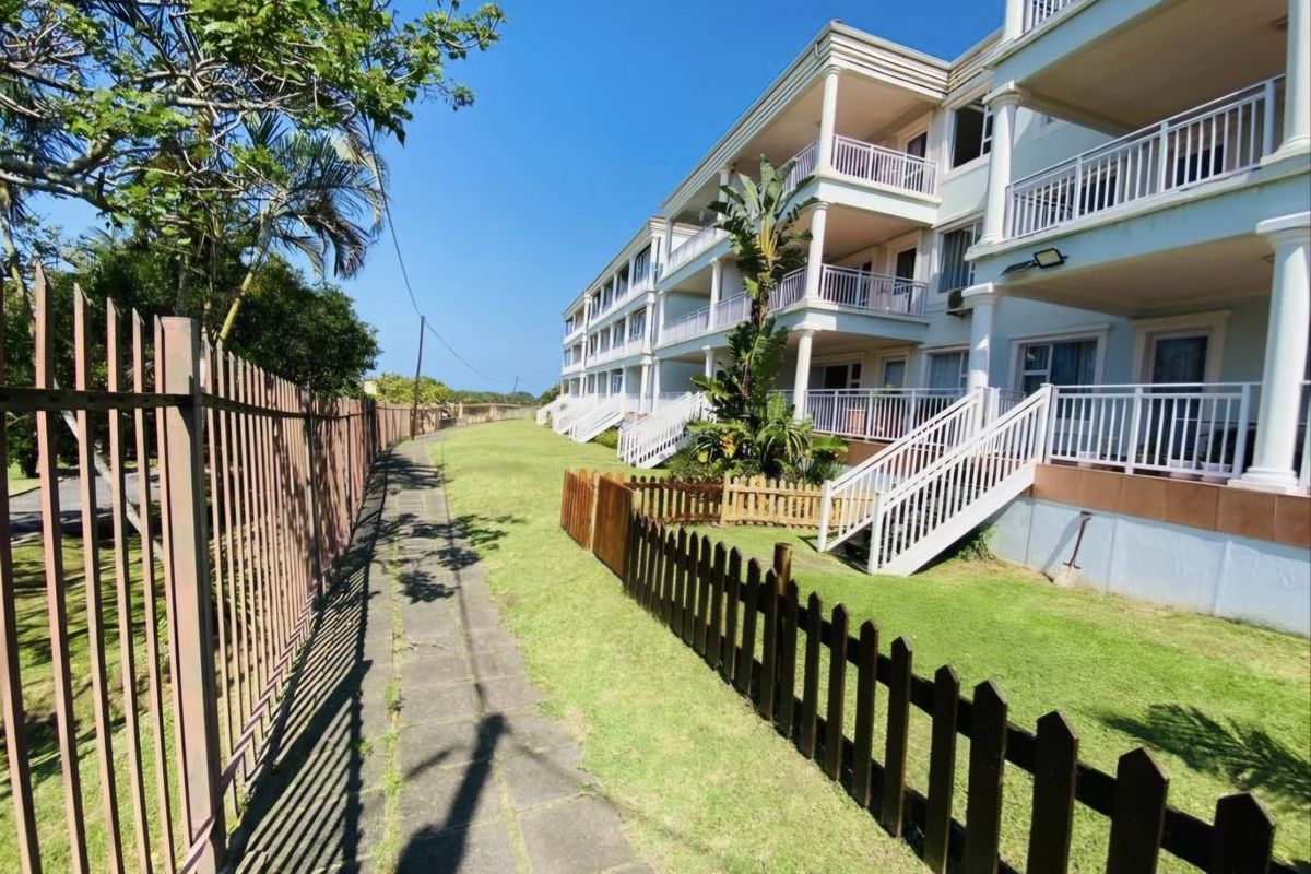 Exterior view of an apartment complex featuring private balconies, external staircases, and a well-maintained lawn area with perimeter fencing.