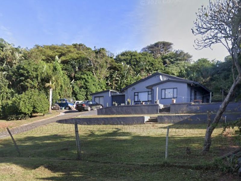Front view of a single-story house featuring a paved driveway, retaining walls, and perimeter security fencing set against a lush, sloped garden backdrop.