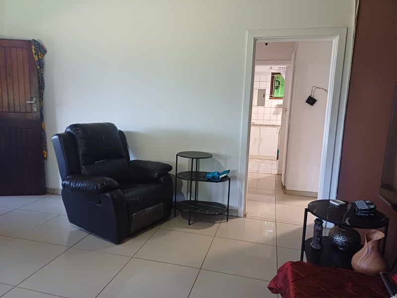 A living room area featuring tiled flooring and a wooden door, with a view through to an adjacent kitchen space.