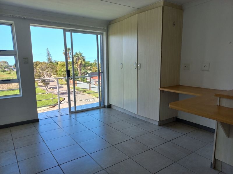 A bright, empty bedroom featuring tiled flooring, built-in cupboards, a built-in desk, and a sliding door leading to a balcony with outdoor views.