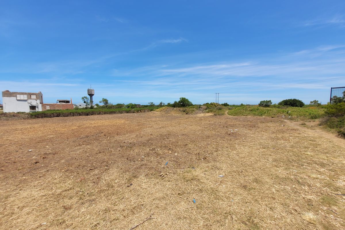 A wide-angle view of a vacant residential plot of land with dry grass and surrounding neighborhood structures in the distance.
