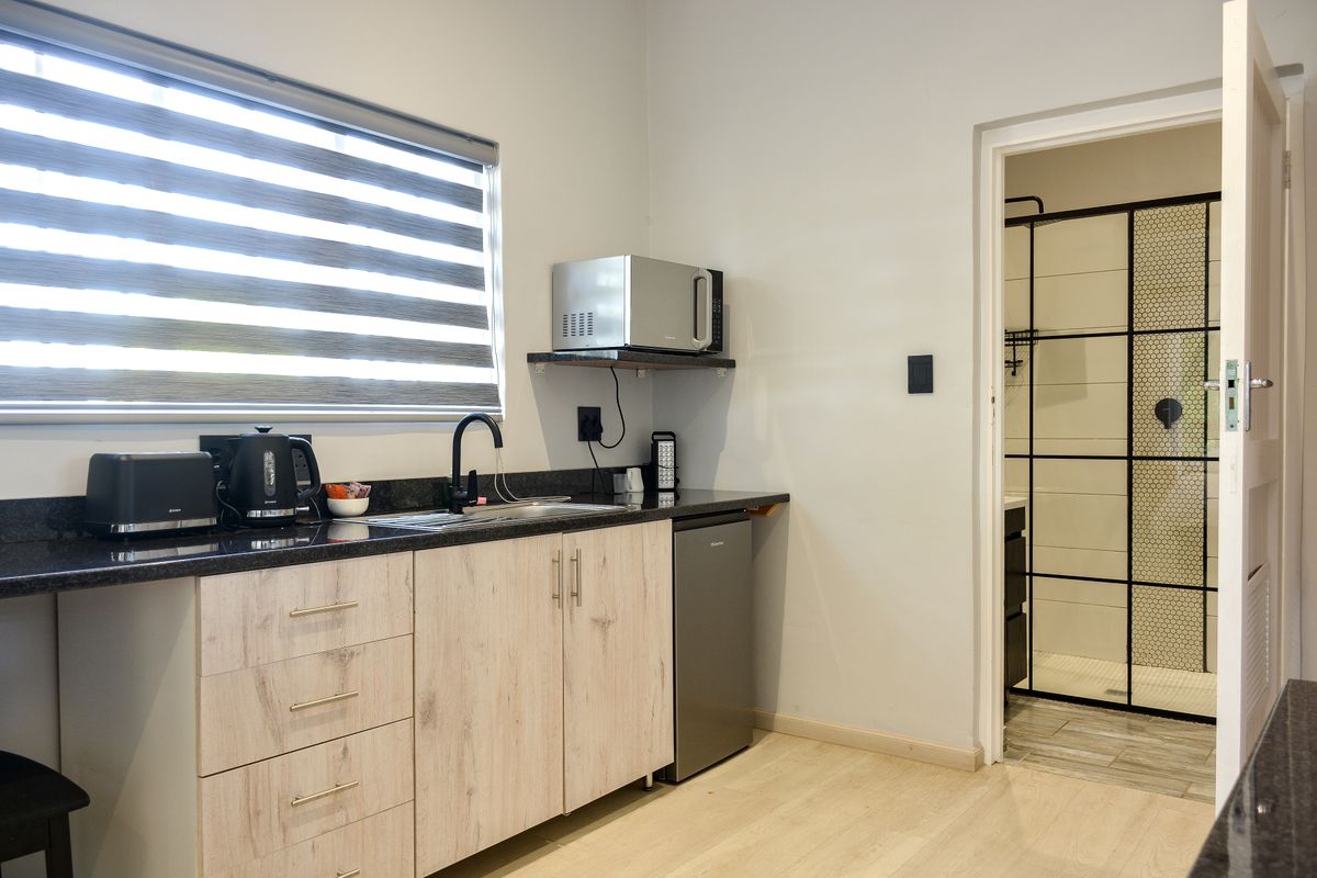 A modern kitchenette featuring light wood-finish cabinetry, granite countertops, and a sleek black tap, with an adjacent bathroom visible through the doorway.