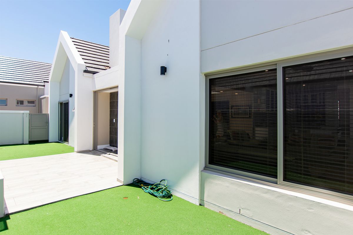 A modern rear exterior view featuring a tiled patio, artificial lawn, and clean white architectural lines with large sliding glass doors.
