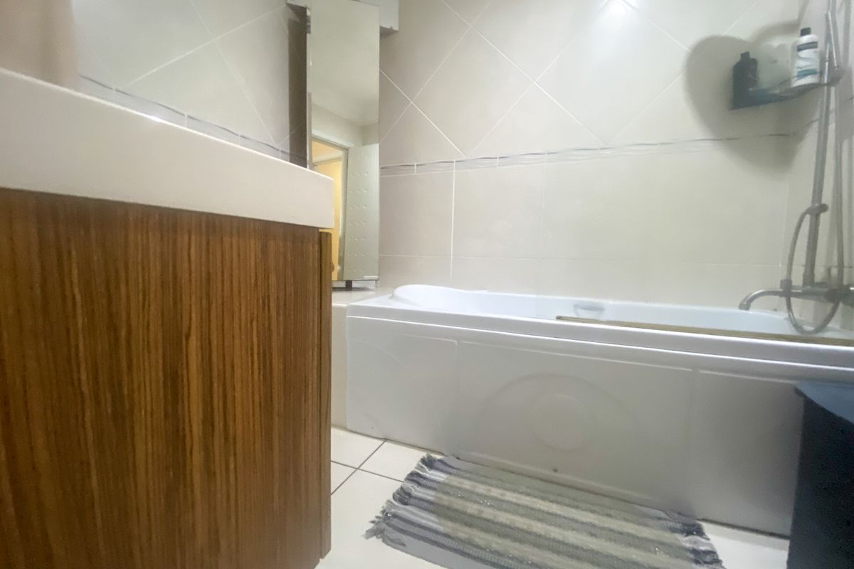 A functional bathroom featuring a built-in bathtub with shower mixer, tiled walls, and a wood-finish vanity unit.