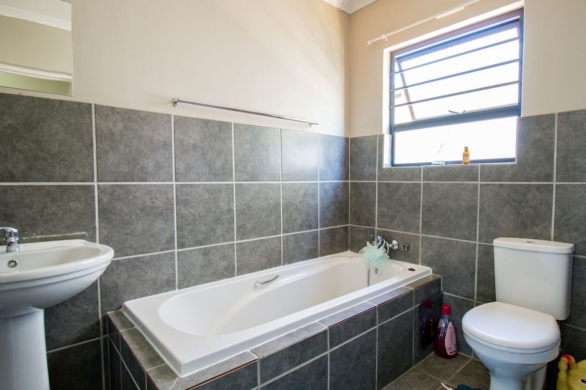 A functional bathroom featuring a pedestal basin, built-in bathtub, and toilet, finished with grey wall and floor tiles and a louver window.