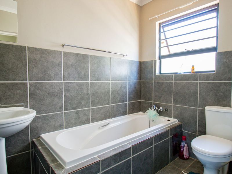 A functional bathroom featuring a pedestal basin, built-in bathtub, and toilet, finished with grey wall and floor tiles and a louver window.