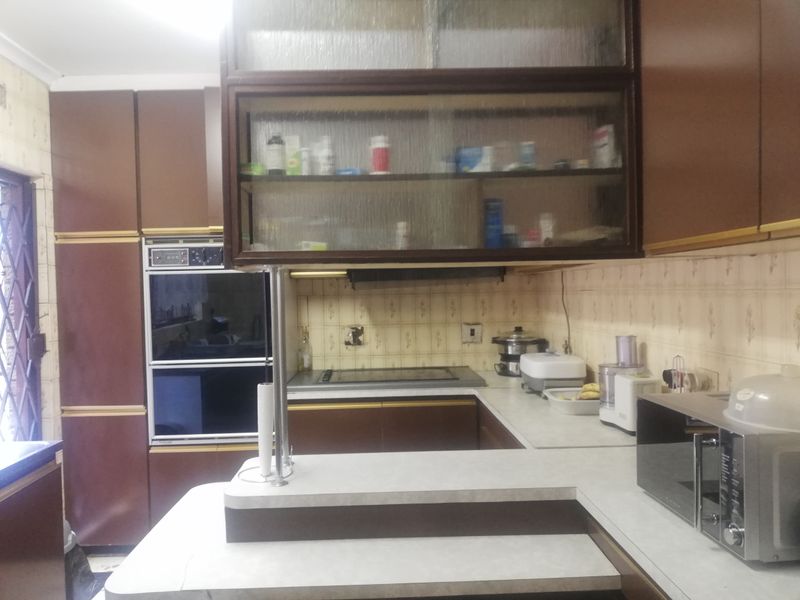 A dated kitchen featuring built-in double ovens, a hob, tiled backsplash, and extensive brown cabinetry with glass-fronted display units.